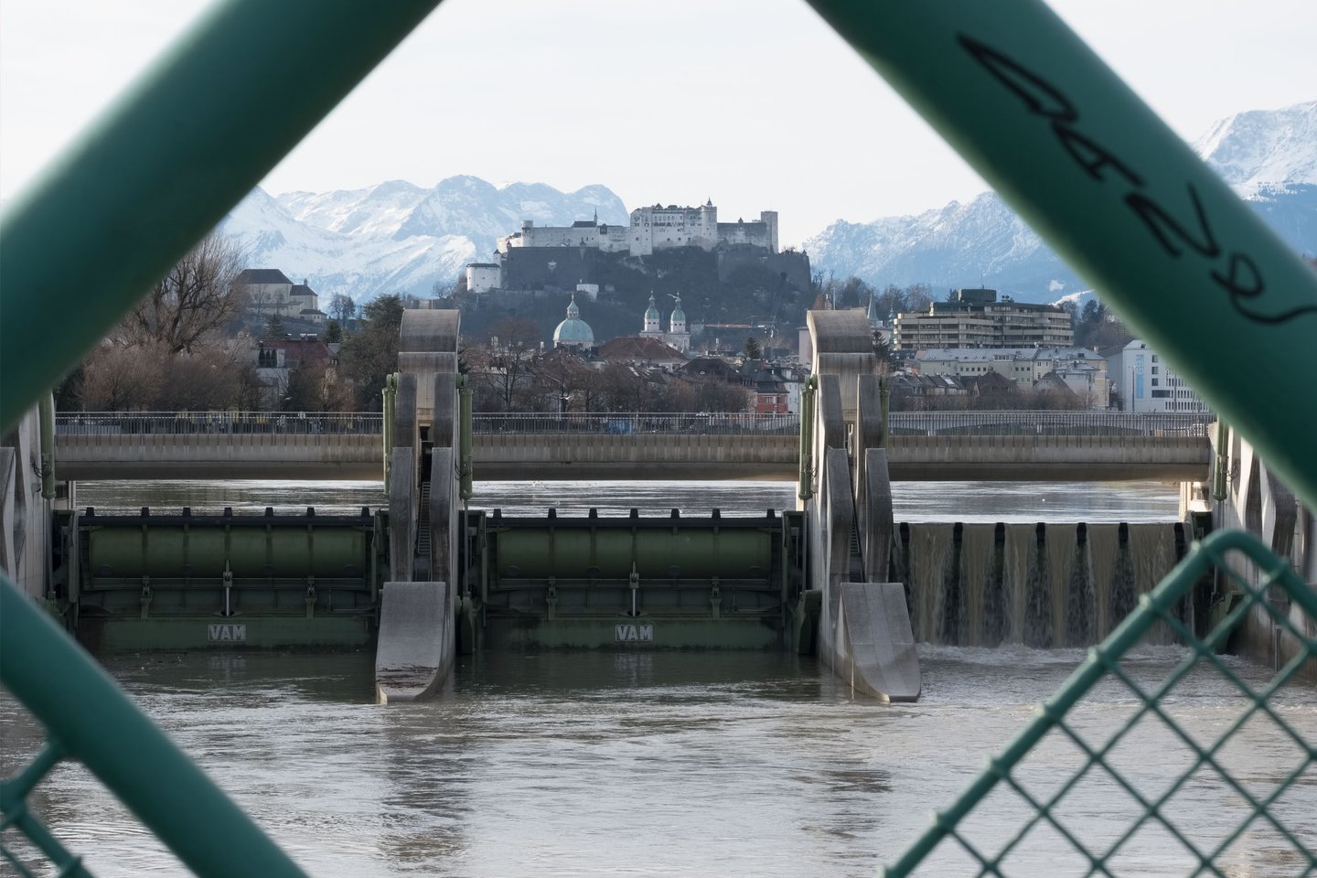 View of Salzach weir through a diamond shaped fence with Hohensalzburg Fortress beyond in Salzburg.