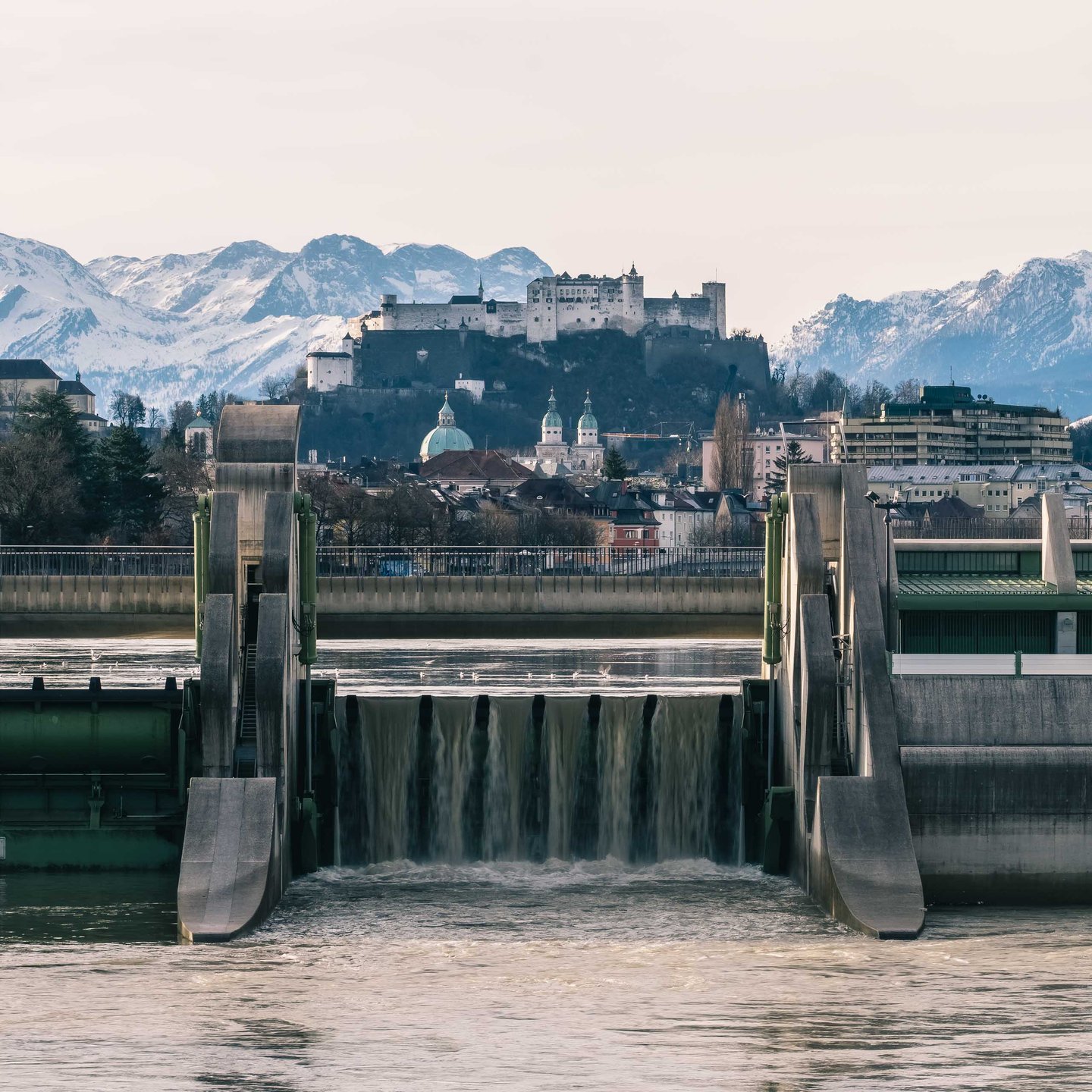 Salzach river weir and waterfall in foreground with Hohensalzburg Fortress and snowy Alps behind.