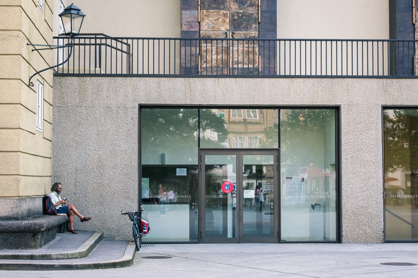 A man sits alone on a stone step beside a glass festival entrance lettered SALZBURGER FESTSPIELE, where a poster announces 100 Jahre Salzburger Festspiele, 1 to 30 August 2020.