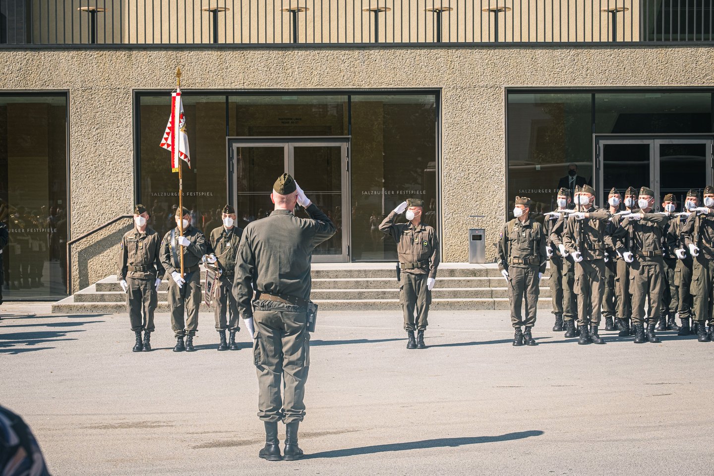 An officer salutes a drawn-up honour guard outside the Festspielhaus entrance, a standard bearer with the red and white Salzburg flag at his left and a rank of soldiers presenting arms at right.