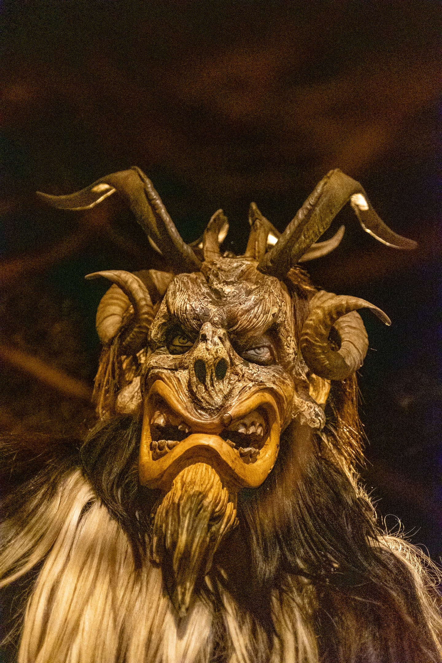 A close portrait of a carved wooden Krampus mask with curled and straight horns, bared teeth, a hooked wooden beard and pale eyes, lit warm against the black night.