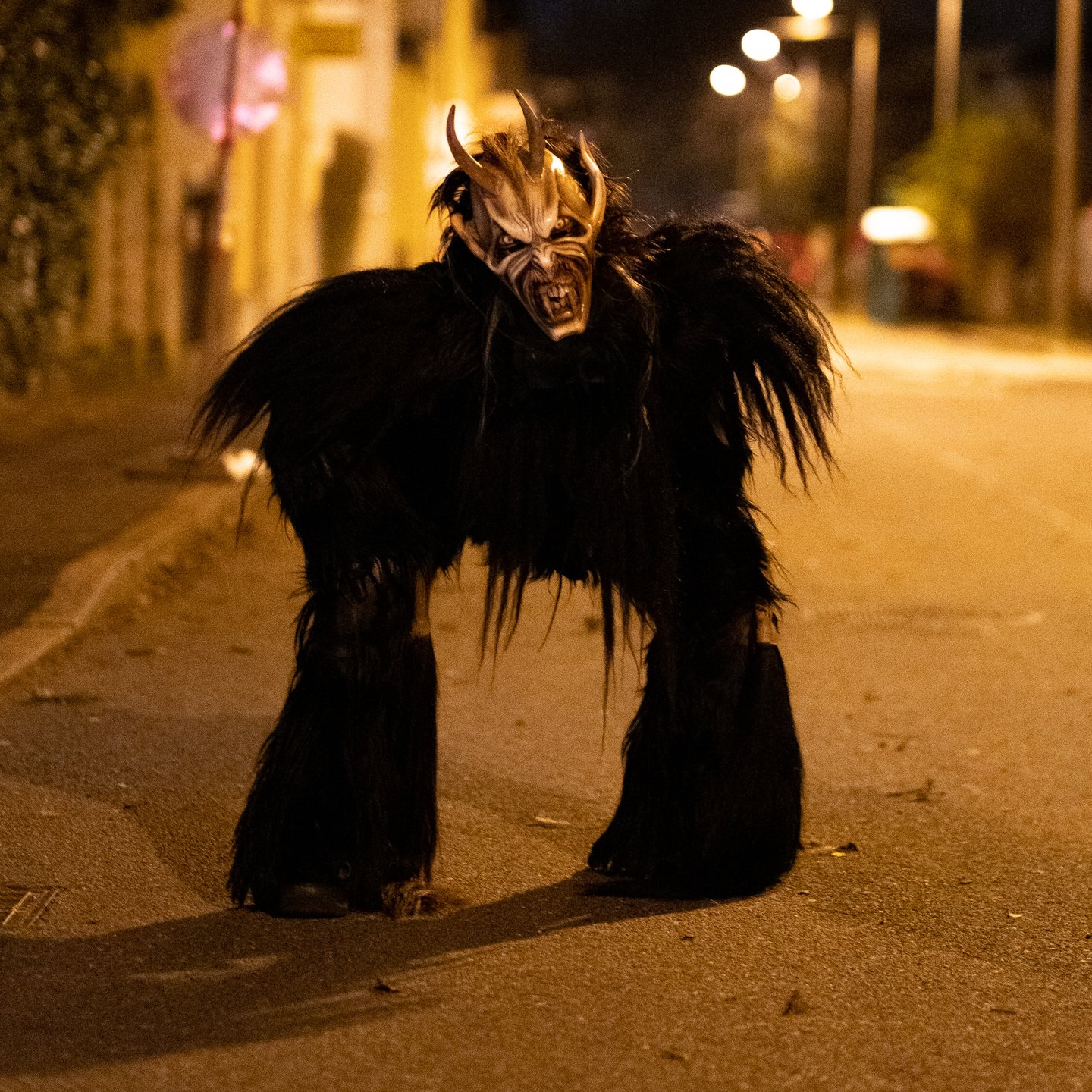 A single Krampus in black fur with a pale carved mask and swept back horns stands alone in the middle of an empty village street washed orange by sodium lamps.