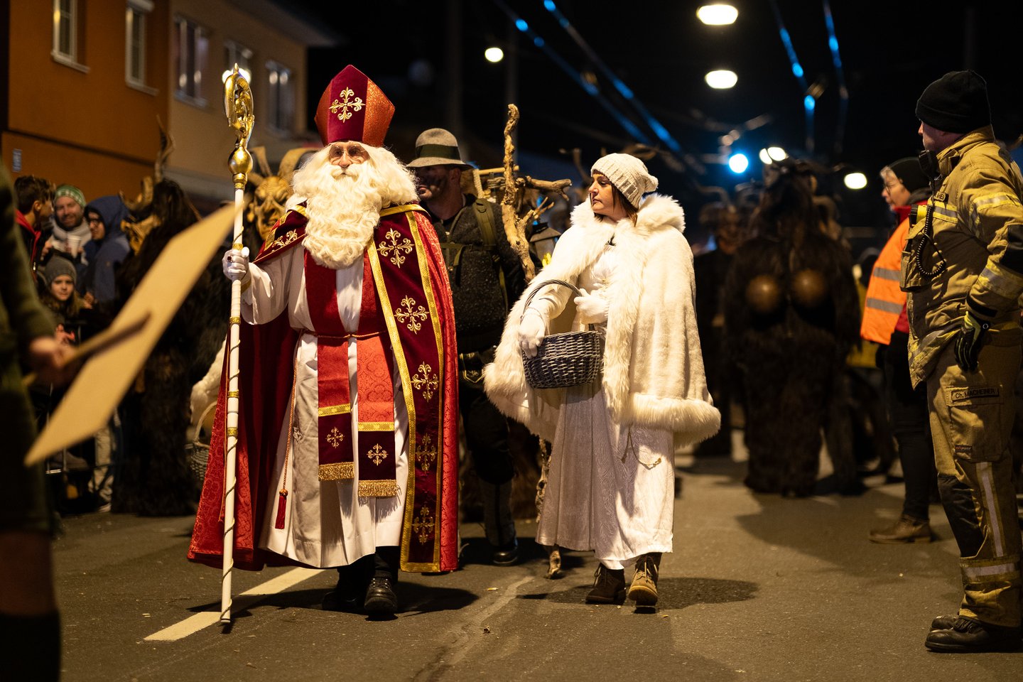 Saint Nicholas in a red and gold cope with a mitre and crozier walks up a village street beside a woman dressed as an angel in a white fur trimmed cape carrying a wicker basket, with masked figures, a lantern bearer and a firefighter behind them.