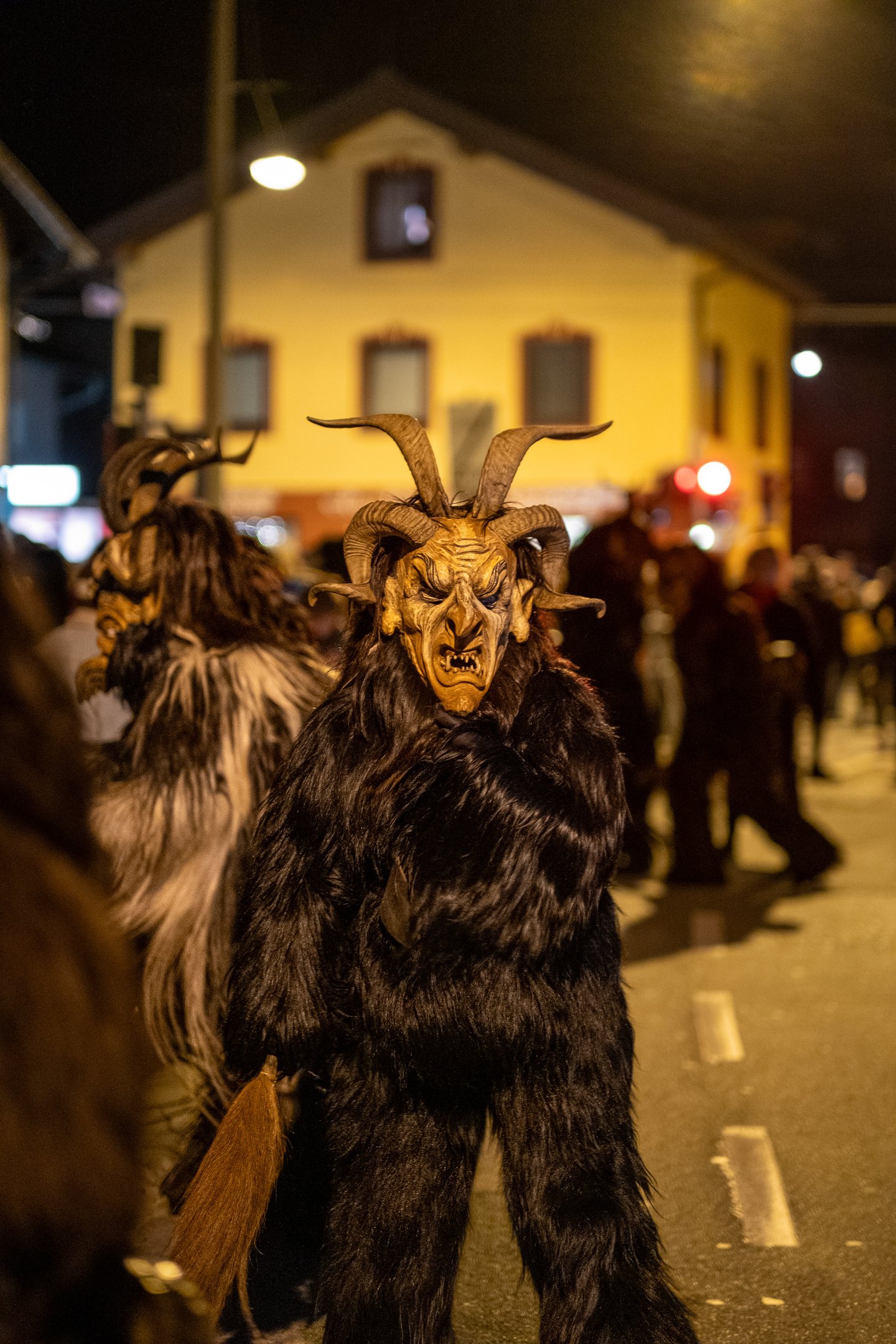 A Krampus in dark shaggy fur wearing a horned wooden mask with curled ram horns and a broom tail stands in the road in front of a yellow gabled village house, with another masked figure beside him.
