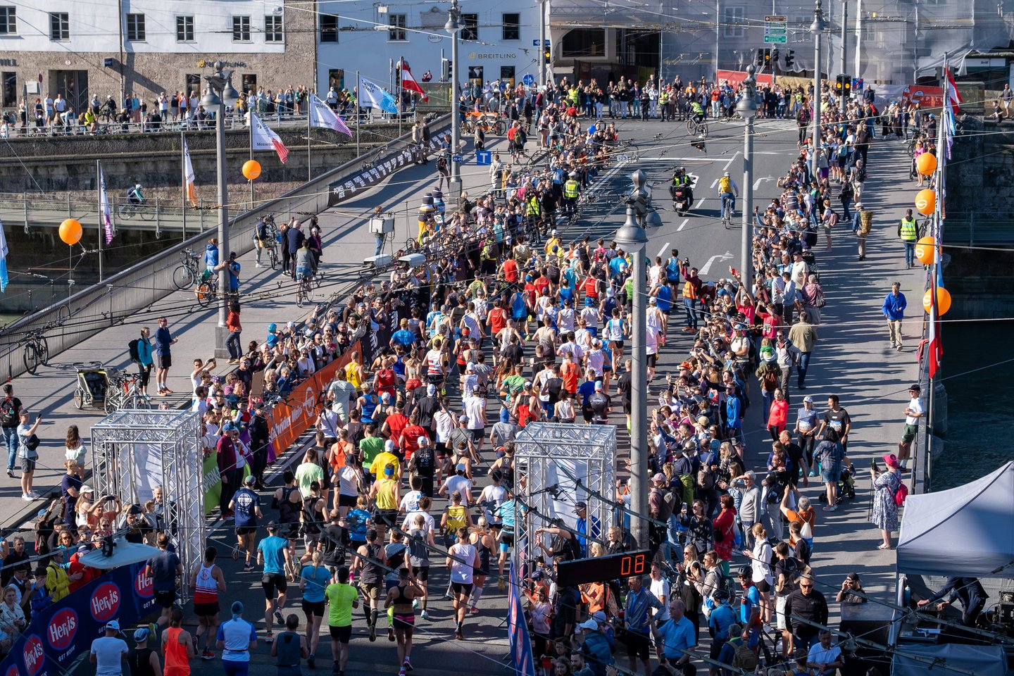The field of runners streams across the Staatsbruecke between orange barriers and cheering spectators, passing the race clock and the festival flags on the bridge.
