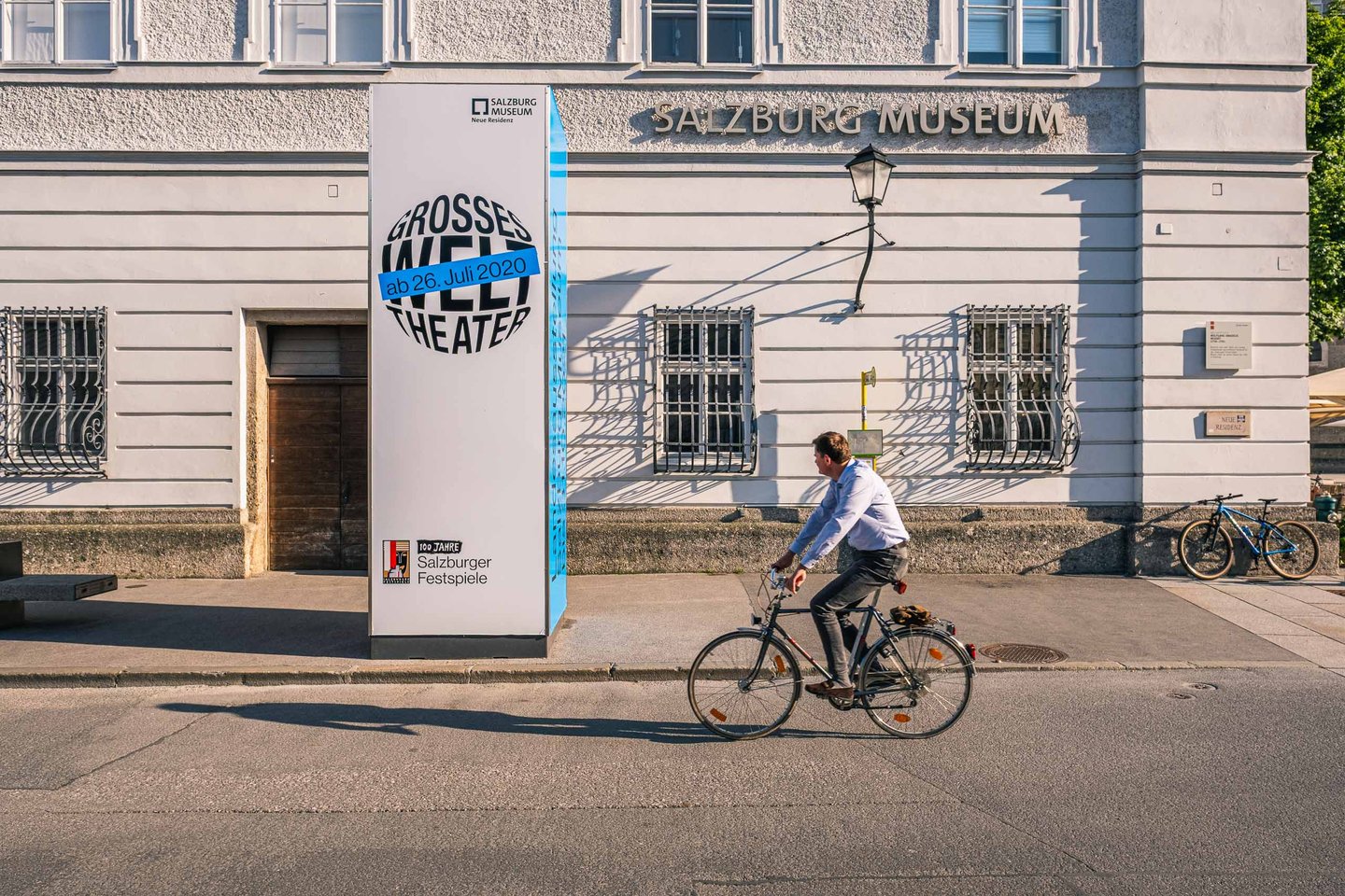 Cyclist passing Salzburg Museum poster column in front of Neue Residenz facade in Salzburg old town