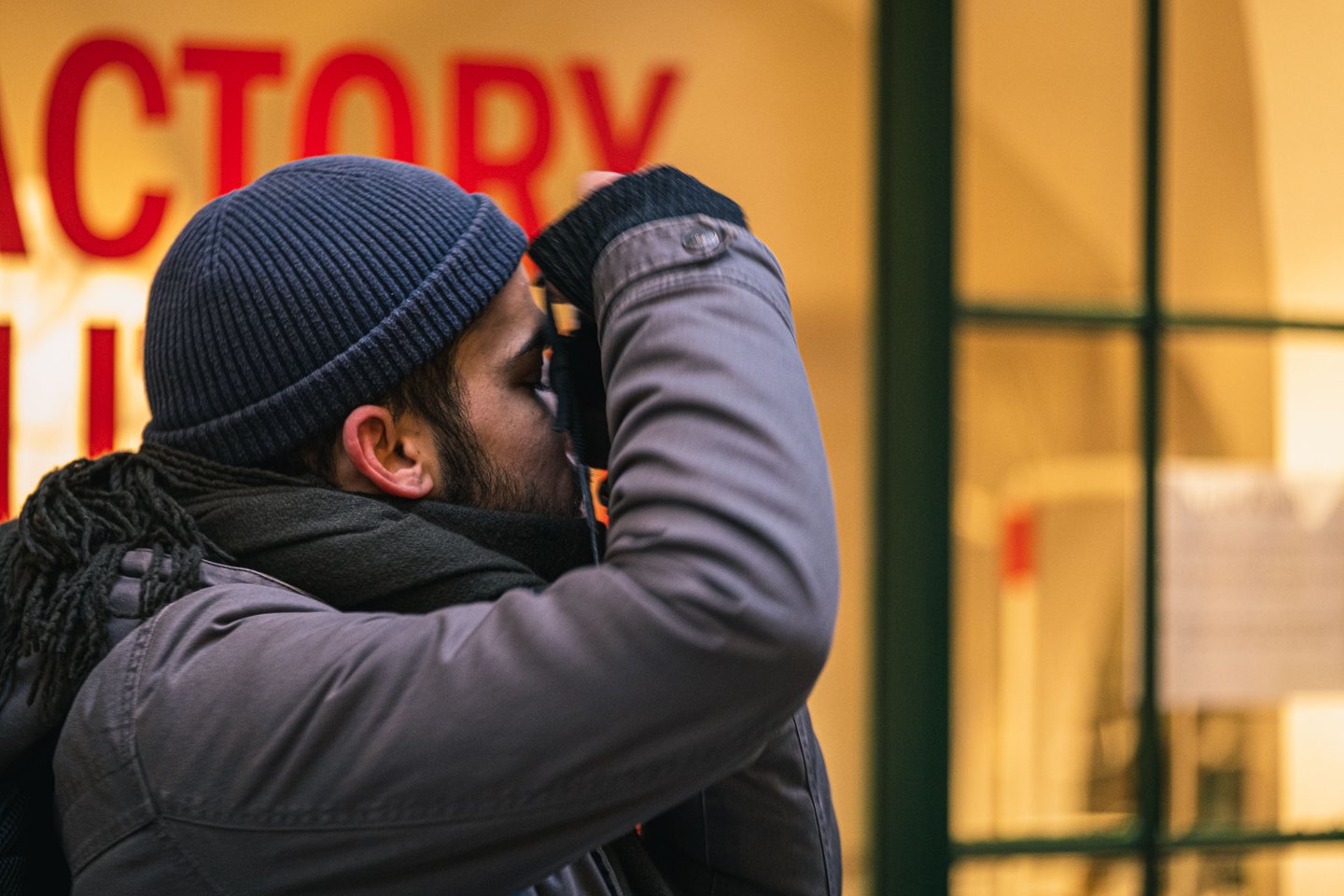 Close-up portrait of a travel photographer in beanie and scarf with a red FACTORY shop sign behind used as content asset for photography articles