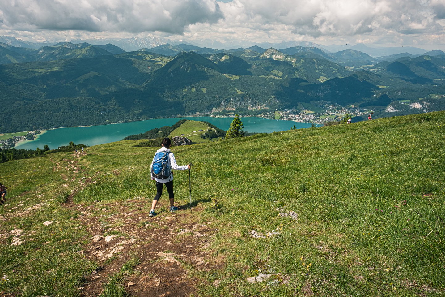A hiker with a blue backpack and a walking pole heads down the Schafberg meadow path toward the turquoise Wolfgangsee, with St. Wolfgang spread along the far shore.