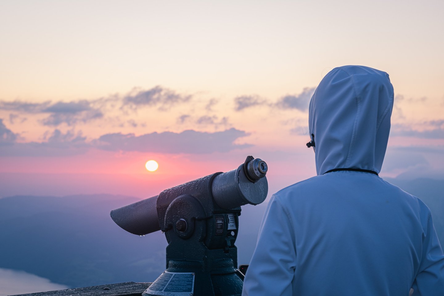 A dew-covered coin telescope points at the rising sun from the Schafberg summit while a person in a white hooded jacket stands beside it, watching the light come up over the haze.