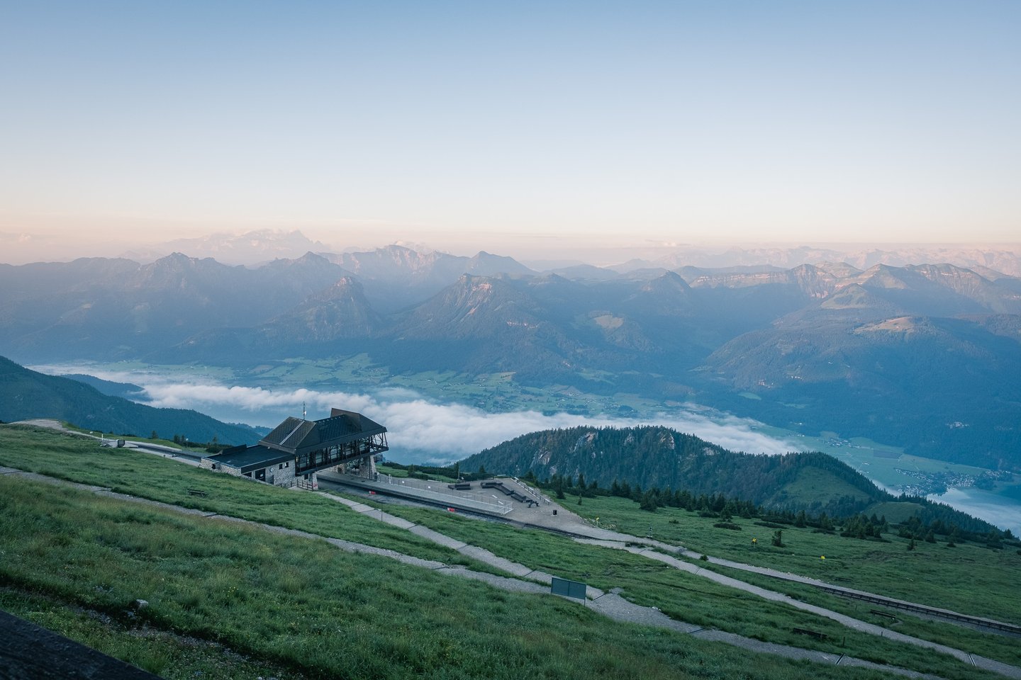 A broad dawn panorama from the Schafberg summit takes in the railway station on its shoulder, the fog-filled Wolfgangsee basin and range upon range of Salzkammergut peaks under an orange horizon.