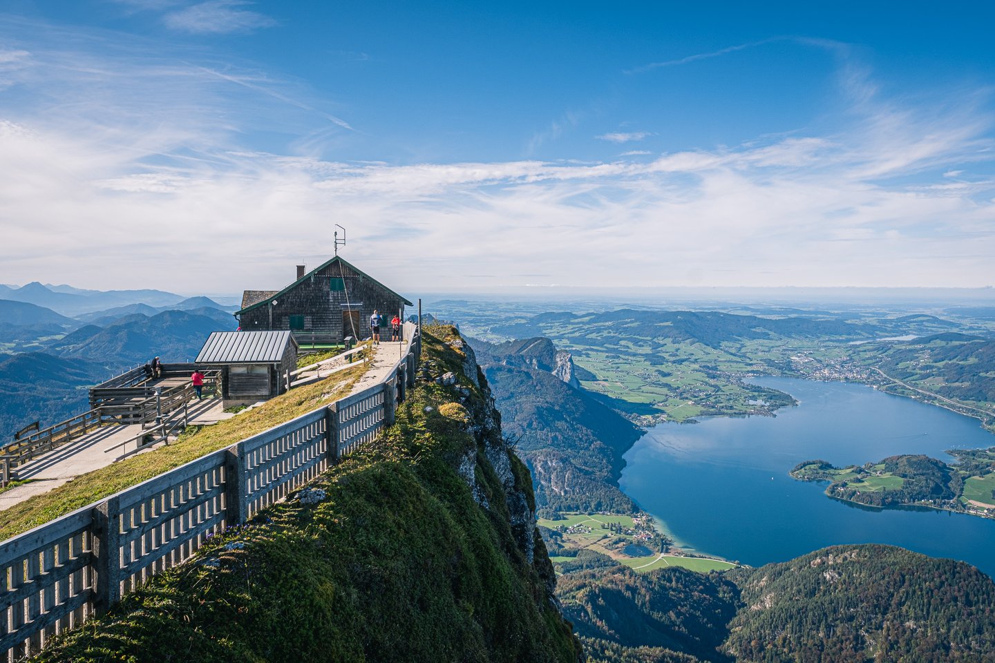 The shingled Himmelspforte hut stands on the narrow Schafberg summit ridge, with a concrete walkway and slatted railing running along the cliff edge high above the Mondsee.