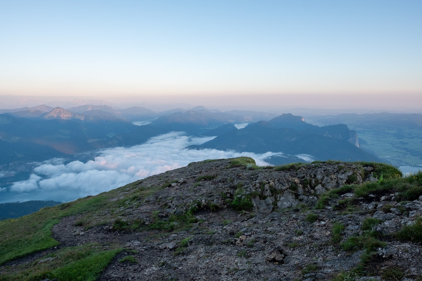The bare limestone crest of the Schafberg summit falls away into a thick cloud inversion filling the valleys, with a second lake and a pale cliff face showing beyond in the early morning haze.