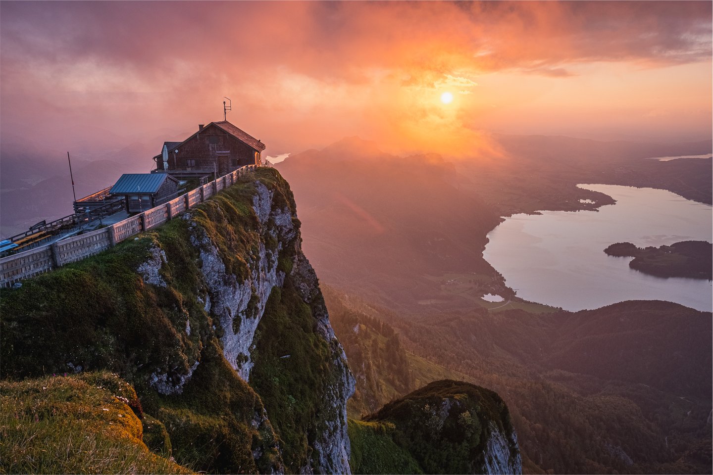 From the Schafberg cliff edge beside the summit house, the sun sinks through pink and orange cloud over the Mondsee, whose pale water and wooded peninsula fill the valley far below.