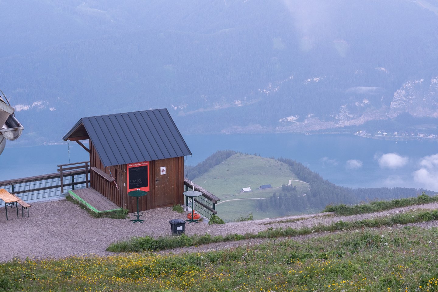 A small wooden kiosk hut with a red chalkboard menu stands on the gravel terrace at the Schafberg summit, high above the blue Wolfgangsee, a green alm knoll and the steep wooded far shore.