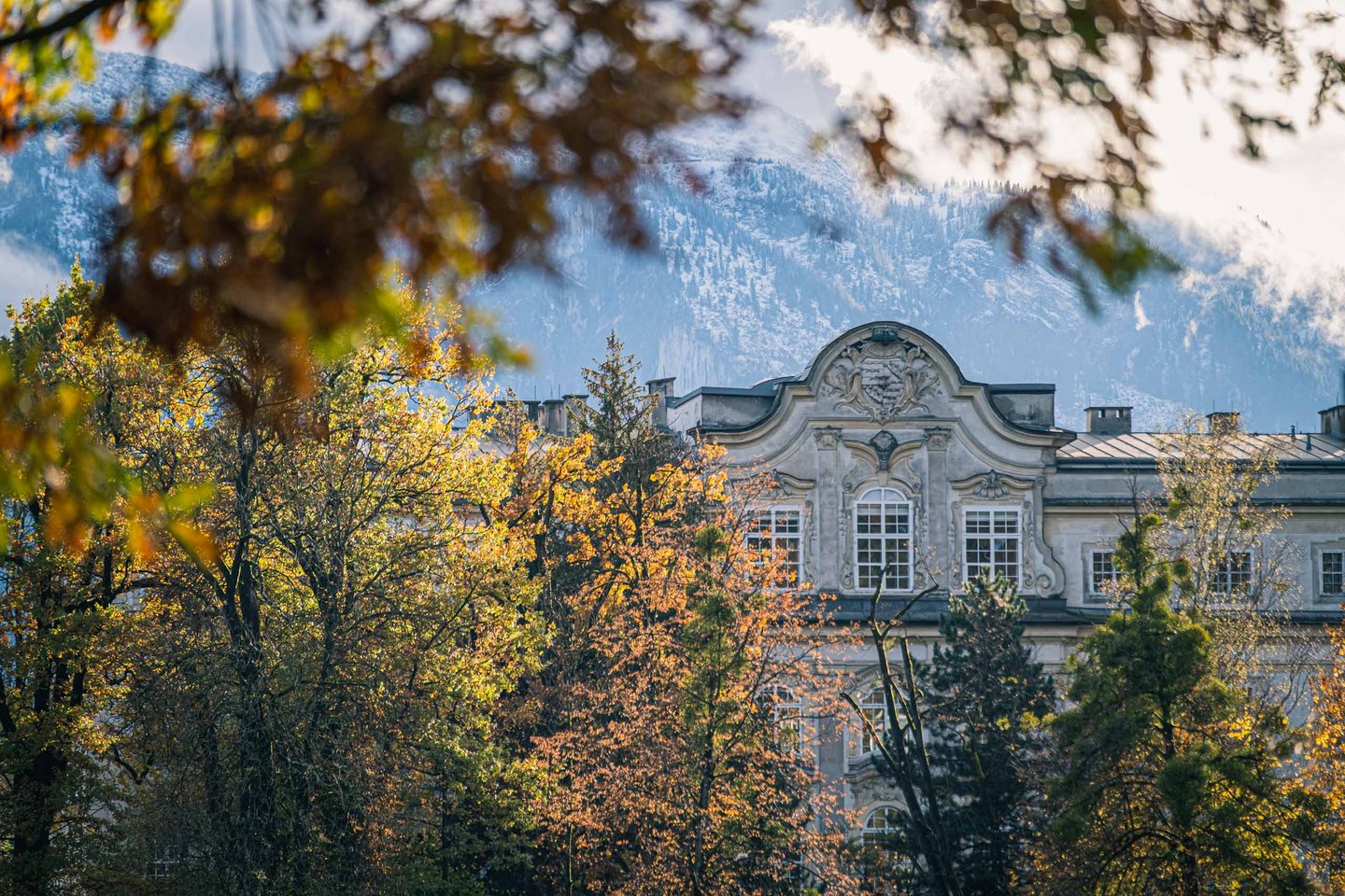 Ornate baroque facade gable of Schloss Leopoldskron framed by autumn trees with mountains behind in Salzburg.