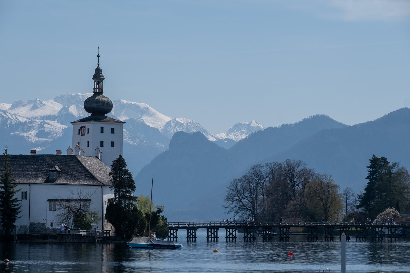 The onion-domed clock tower of Seeschloss Ort rises above the Traunsee, with the long wooden footbridge to the island and snow-covered mountains behind.
