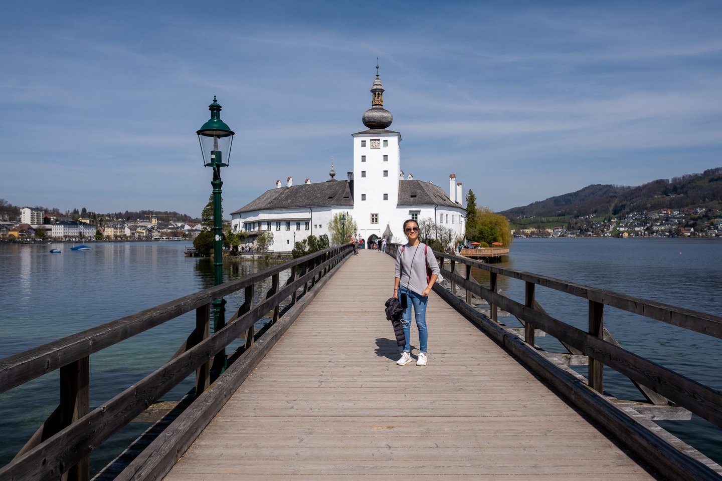 A visitor stands on the long wooden footbridge leading to Seeschloss Ort, a green cast-iron lamp post beside her and the castle's white clock tower straight ahead.