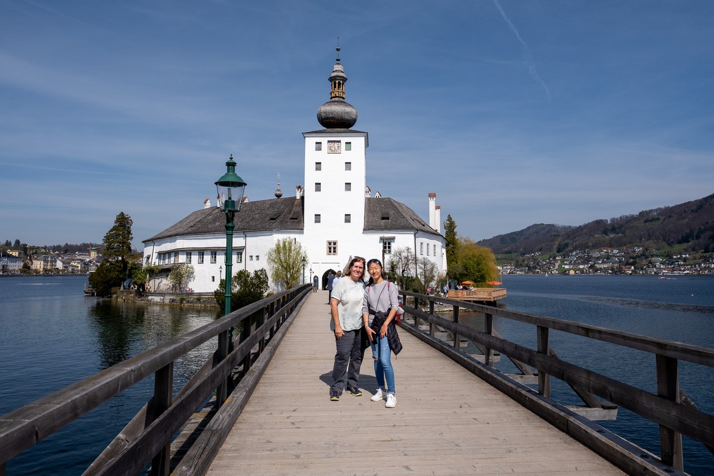 Two women stand side by side on the wooden footbridge to Seeschloss Ort, with the castle, the Traunsee and the hillside houses of Gmunden behind them.