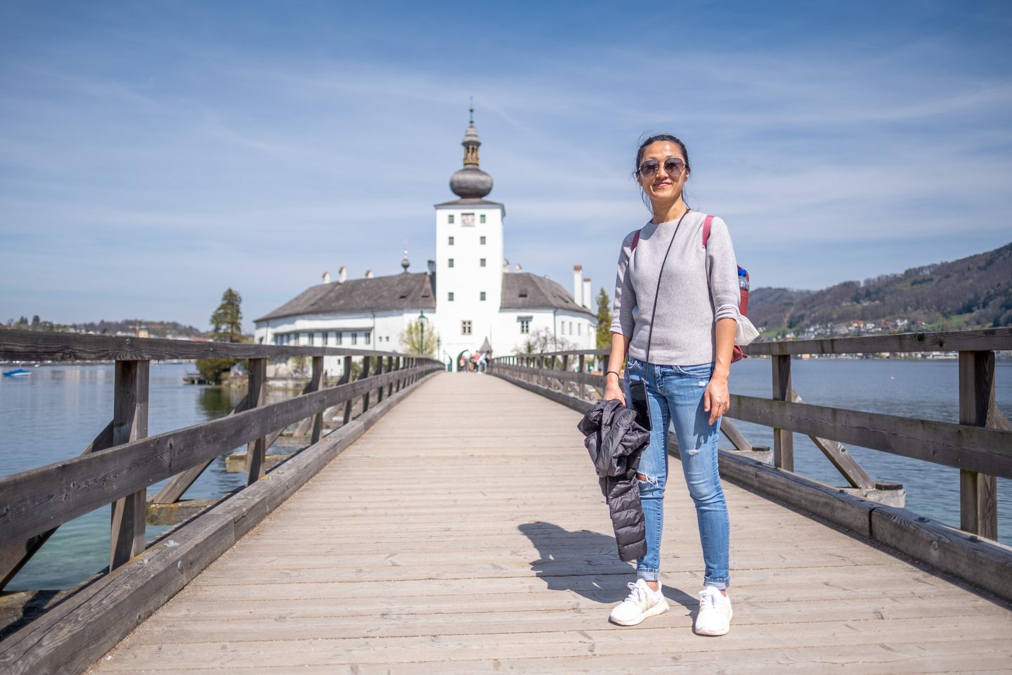 A woman in a grey jumper and sunglasses stands on the wooden footbridge to Seeschloss Ort, the white castle and its onion dome behind her.