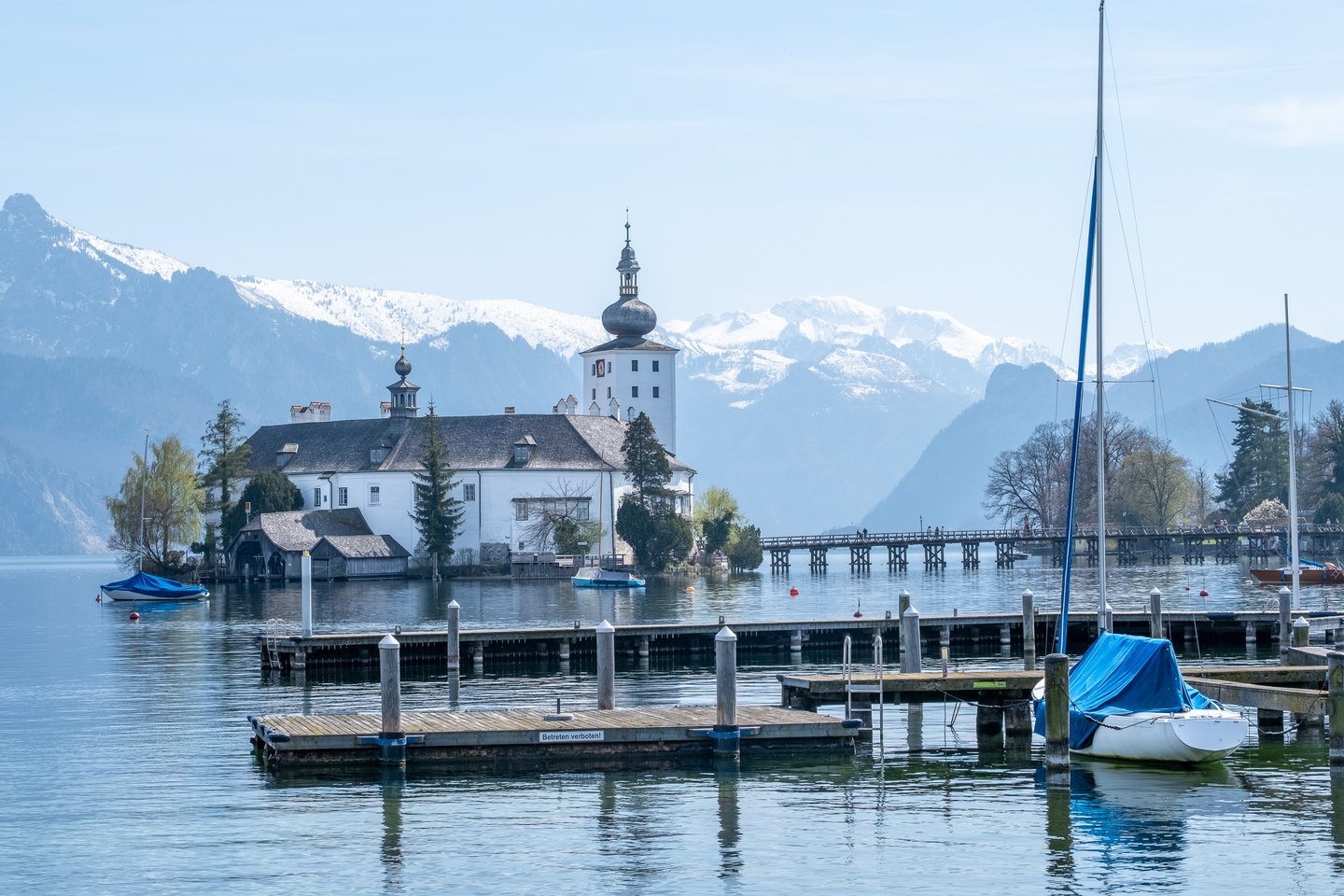 Seeschloss Ort on its island in the Traunsee seen past a wooden boat jetty with tarpaulin-covered sailboats moored in the foreground.