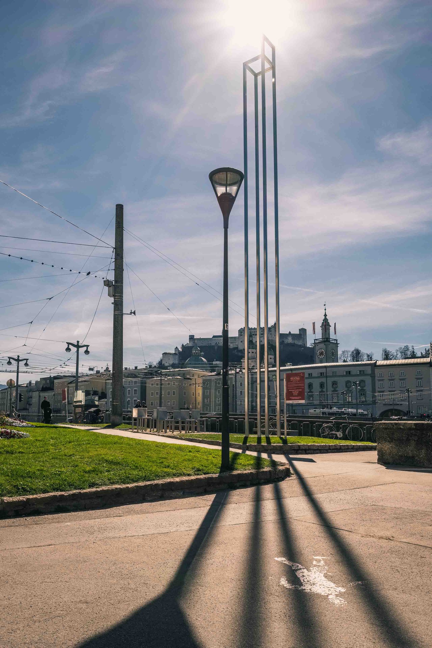 Tall slender open metal frame of the Spirit of Mozart installation on the Salzach embankment with Hohensalzburg Fortress and Glockenspiel visible behind in Salzburg