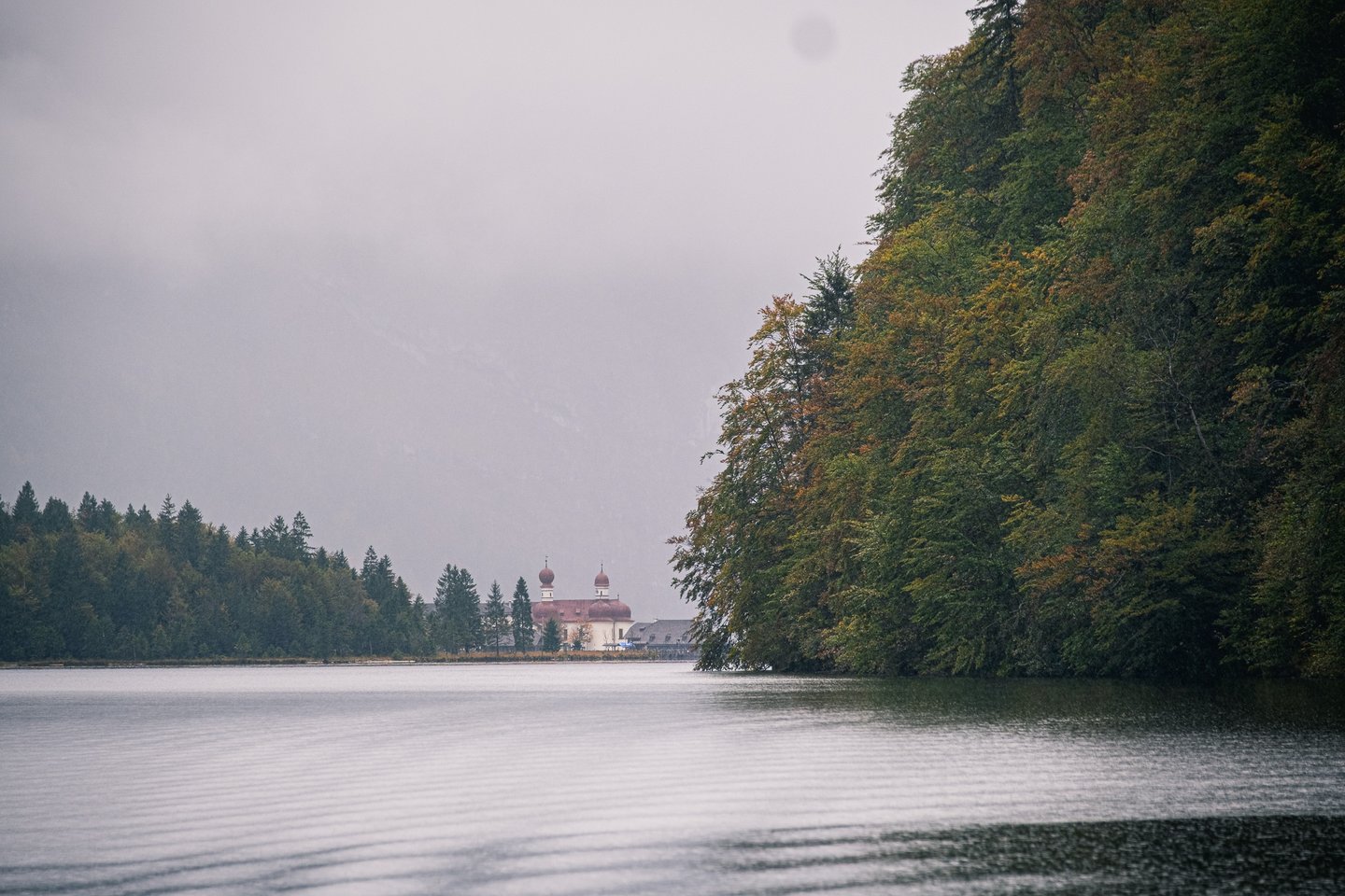 The twin red onion domes of St. Bartholomae chapel show small and pale across the Koenigssee in falling rain, a wooded headland filling the right of the frame.