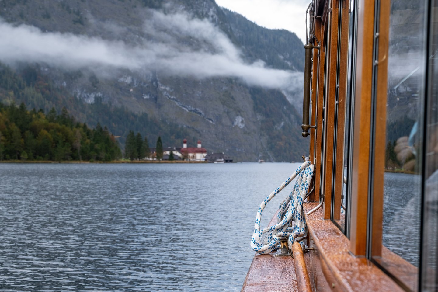 St. Bartholomae with its red onion domes sits on its low green peninsula across the Koenigssee, seen past the varnished rail and coiled mooring rope of the passenger boat.