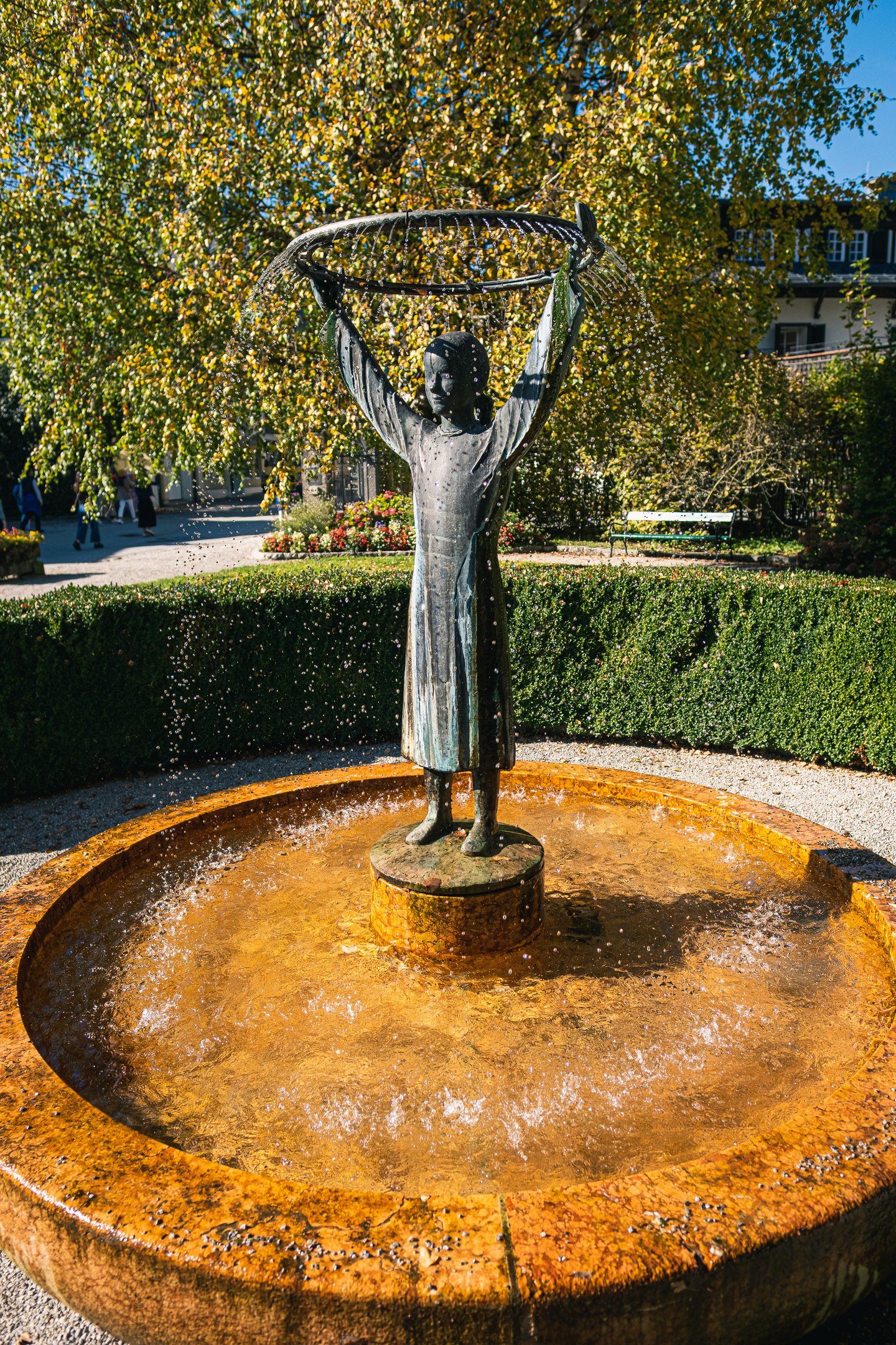 A bronze figure of a girl holds a circular fishing net above her head, water falling from its rim into a round ochre stone basin in a hedged park.