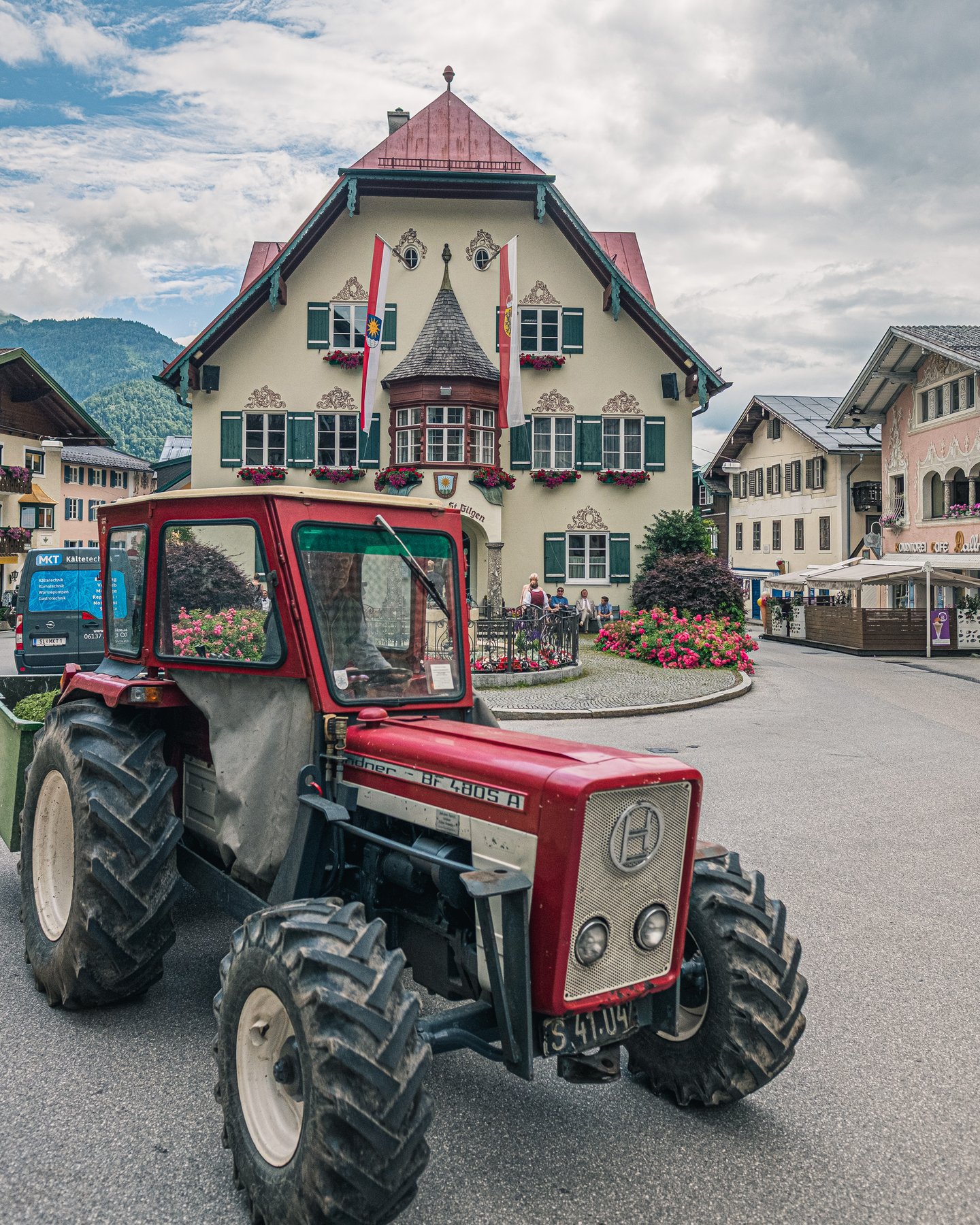 A red Lindner tractor rolls across the square in front of the Rathaus St. Gilgen on a summer day, the building's window boxes full of geraniums and flags hanging from its gable.