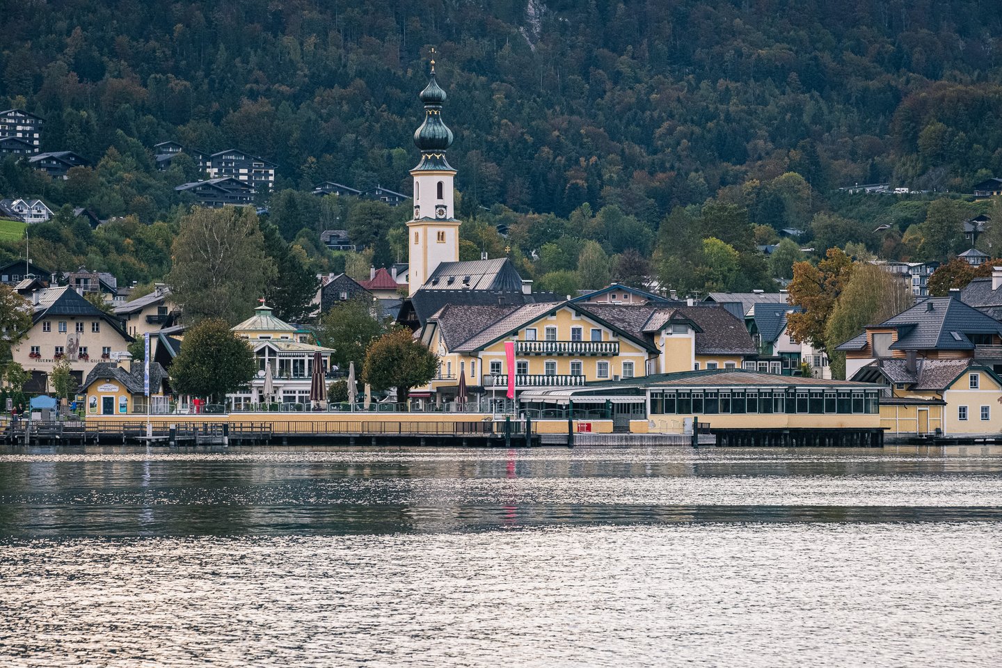 The village of St. Gilgen seen from the water at dusk, its yellow lakefront hotel and glazed terrace below the onion-domed tower of the parish church and a dark wooded slope.