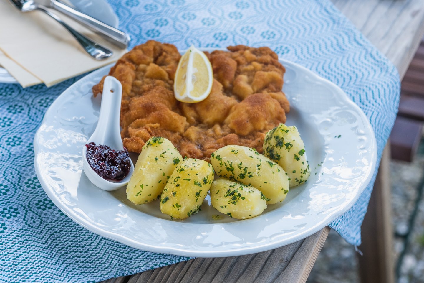 An untouched Wiener Schnitzel with a lemon wedge, parsley potatoes and a spoon of lingonberry jam sits on a white plate at the edge of a weathered wooden outdoor table.