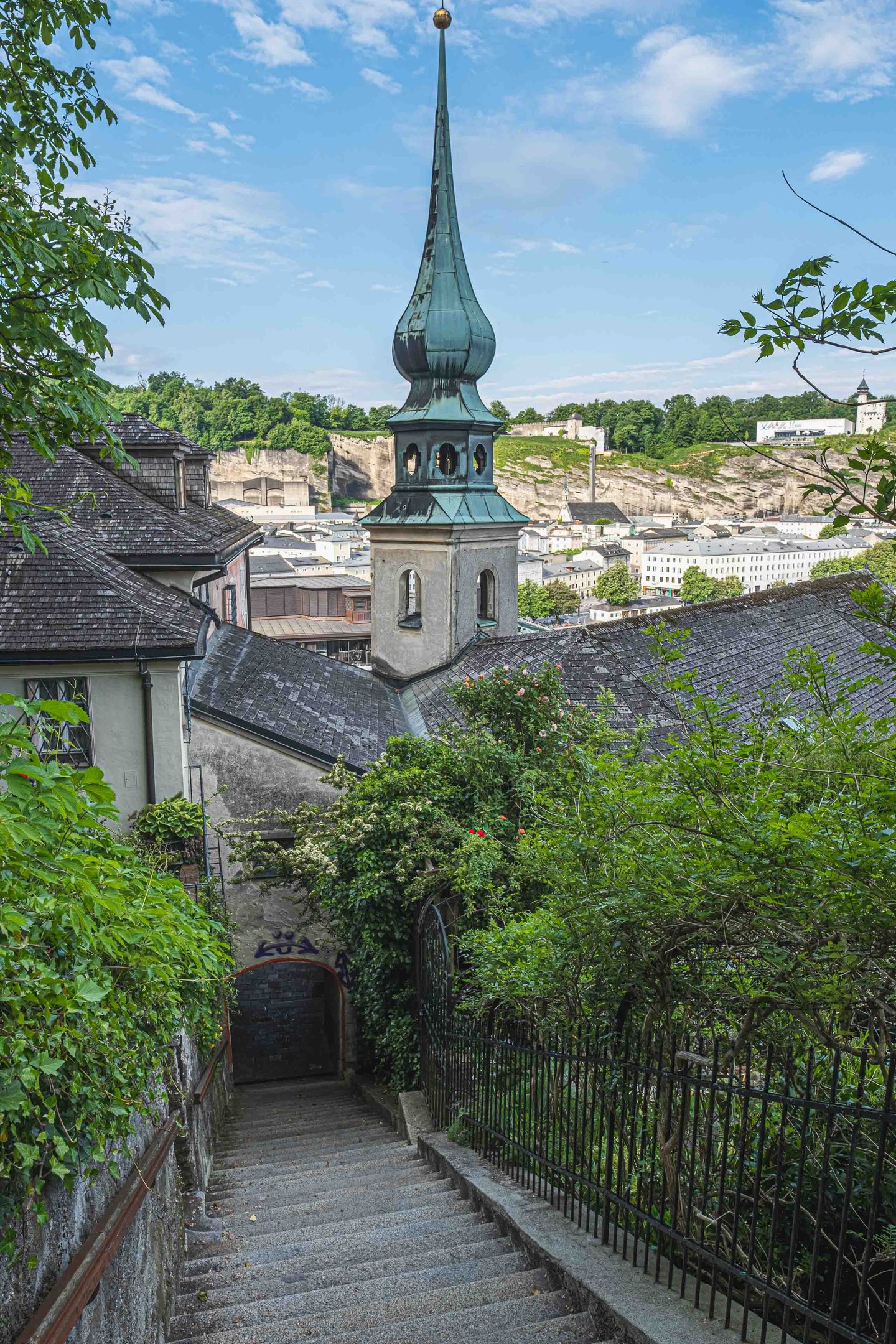 Green onion-dome spire of St John's Church on Imberg seen from stairs descending toward Salzburg old town.