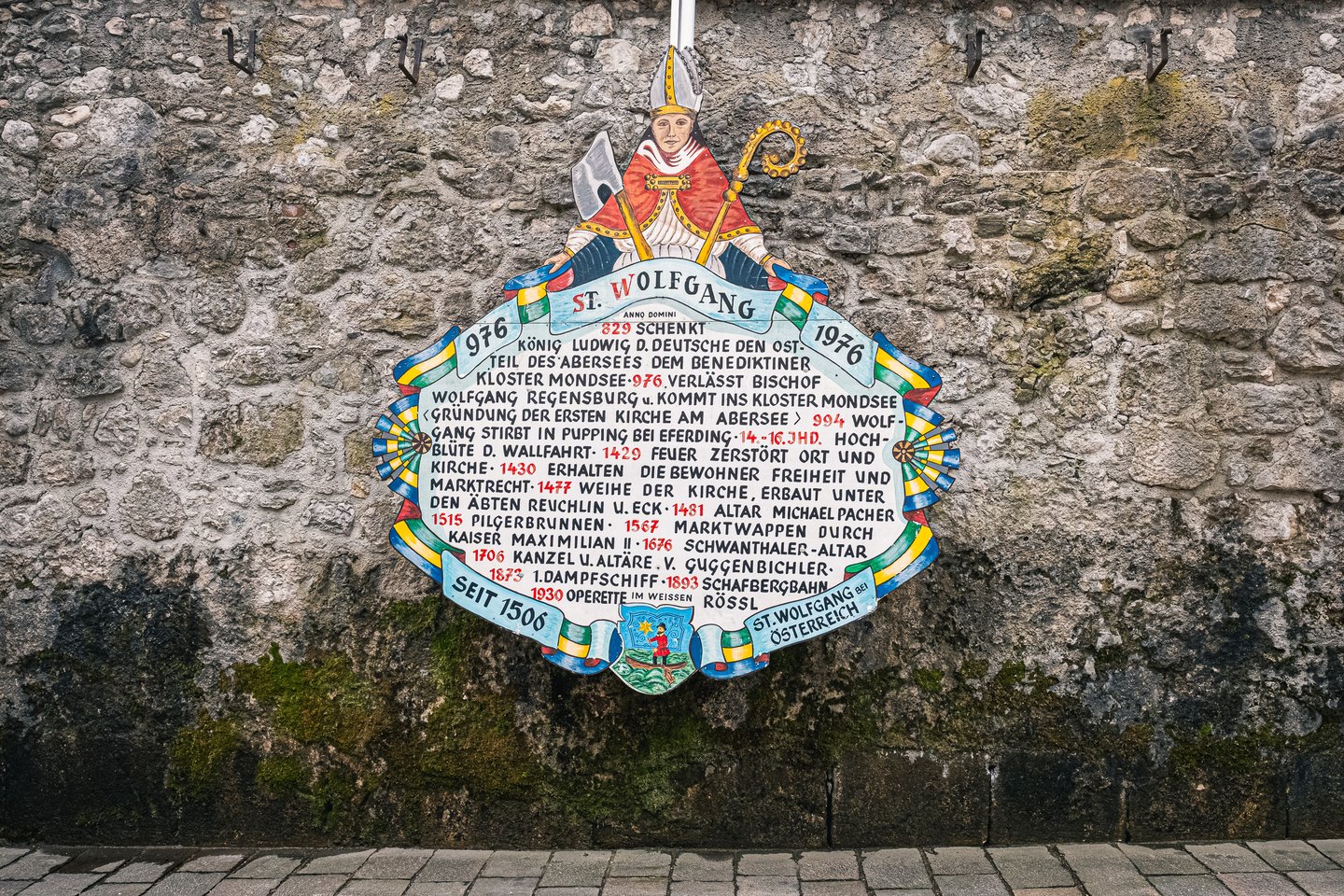 A painted wooden millennium panel on a rough stone wall shows Saint Wolfgang in mitre and red cope holding an axe and crozier above a scrolled chronicle of the village dated 976 to 1976.