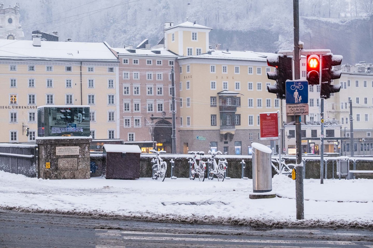 Snow-covered bikes on the Staatsbruecke railing with Altstadt facades and Moenchsberg behind in Salzburg.
