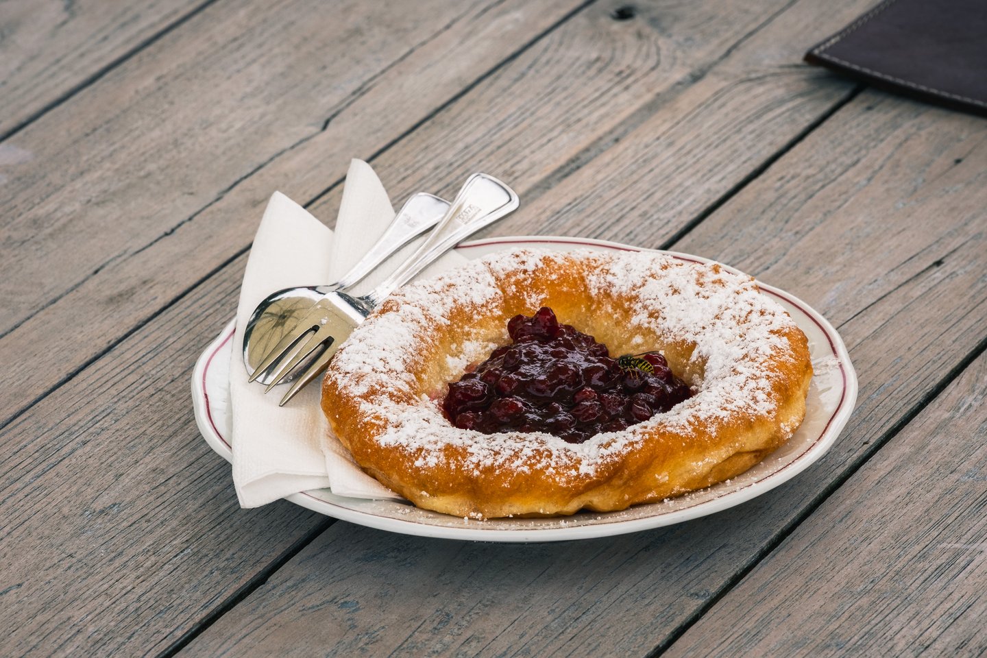 A deep fried Bauernkrapfen dusted with icing sugar and filled with lingonberry jam sits on a red rimmed plate beside a folded napkin and cutlery on a weathered wooden table.