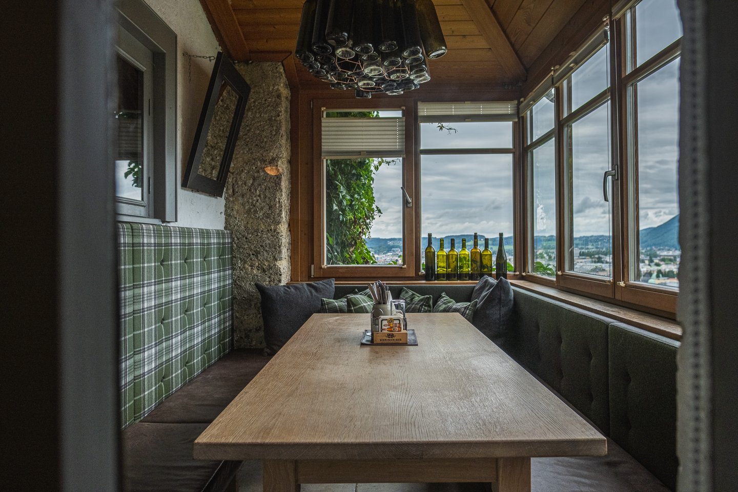 A corner nook inside the Stadtalm on the Mönchsberg with green plaid and dark velvet banquettes around a long oak table, a chandelier made of green wine bottles overhead and the rough conglomerate of the old wall built into the room.