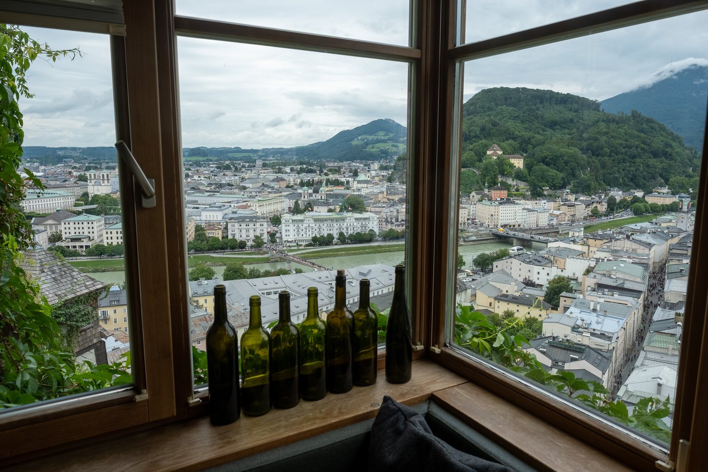Empty green wine bottles line the sill of a corner window in the Stadtalm on the Mönchsberg, looking north over the Salzach and the crowded footbridge to the Kapuzinerberg, with the shingled turret of the old wall just below at the left.