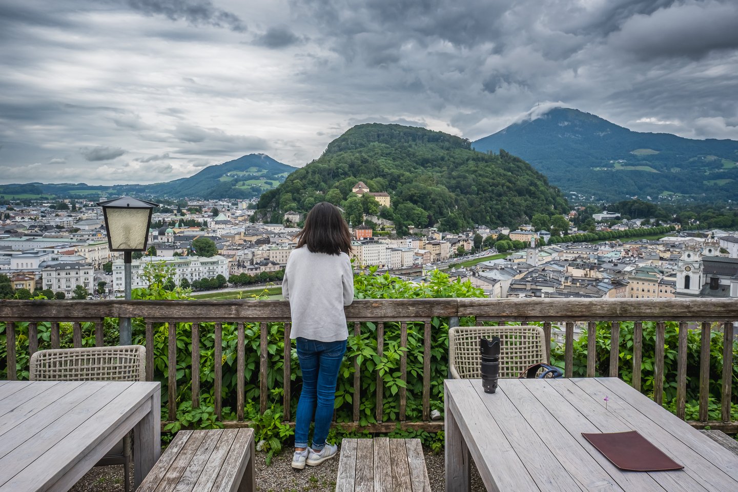 A woman stands with her back to the camera at the wooden railing of the Stadtalm terrace on the Mönchsberg, looking across the Salzburg Neustadt to the Kapuzinerberg with its monastery and the cloud-capped Gaisberg beyond.