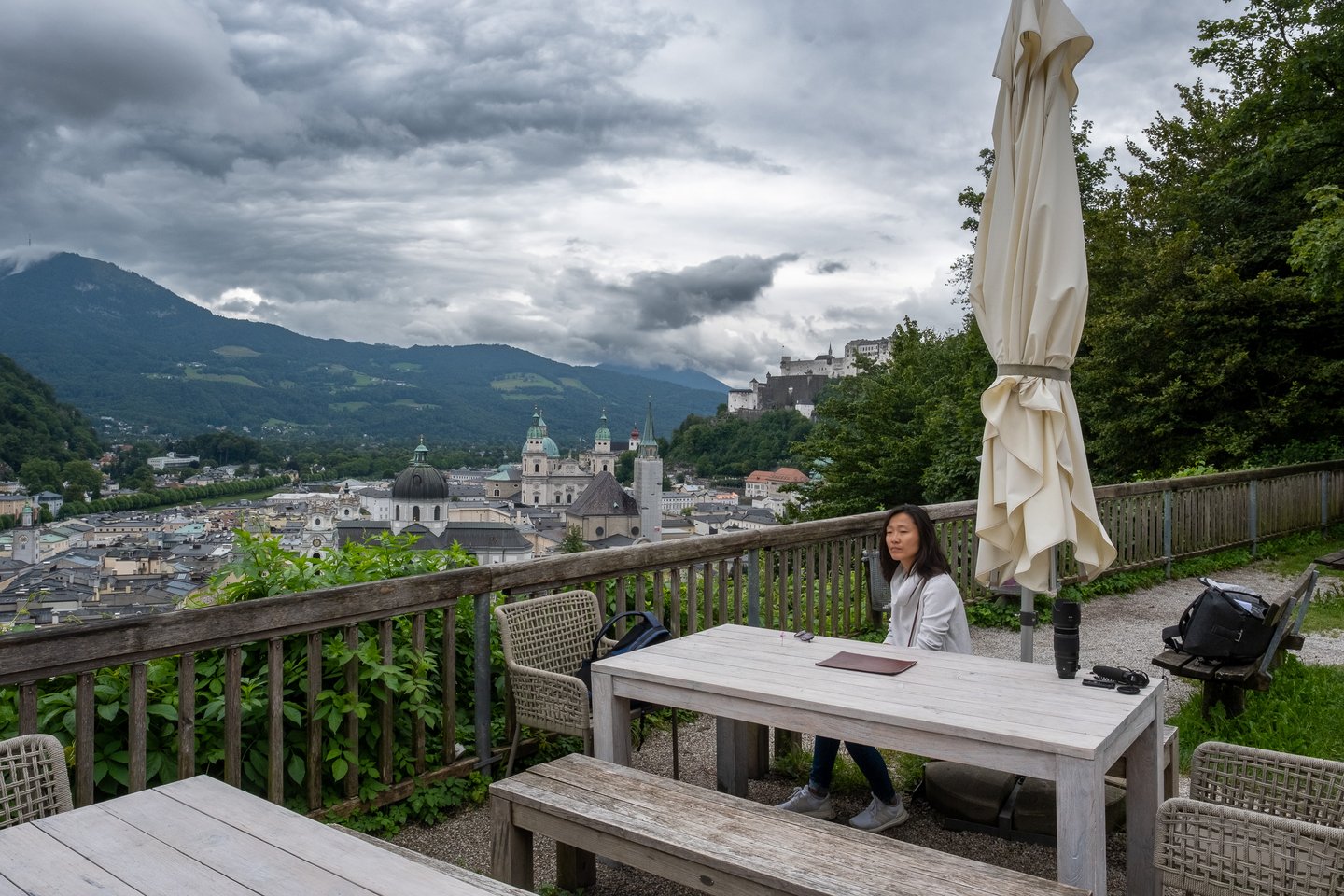 The gravel terrace of the Stadtalm on the Mönchsberg under heavy grey cloud, its weathered tables, benches and a furled umbrella looking out across the Salzburg Altstadt to Hohensalzburg on its ridge.