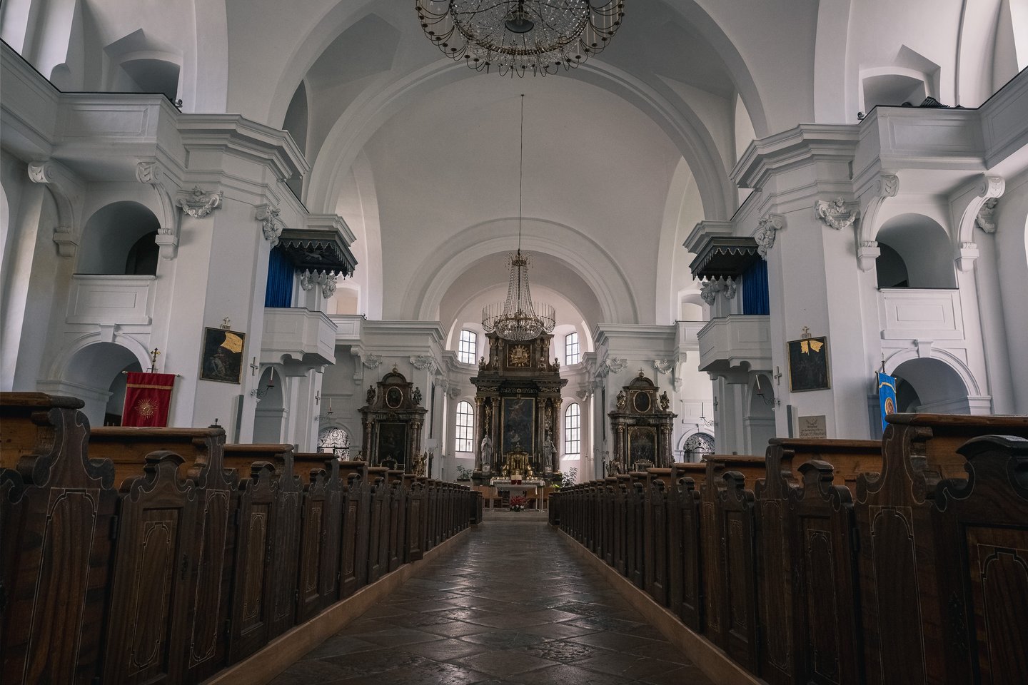 The central aisle of Hallein parish church runs between dark wooden pews toward the altar, under a white barrel vault hung with two crystal chandeliers.
