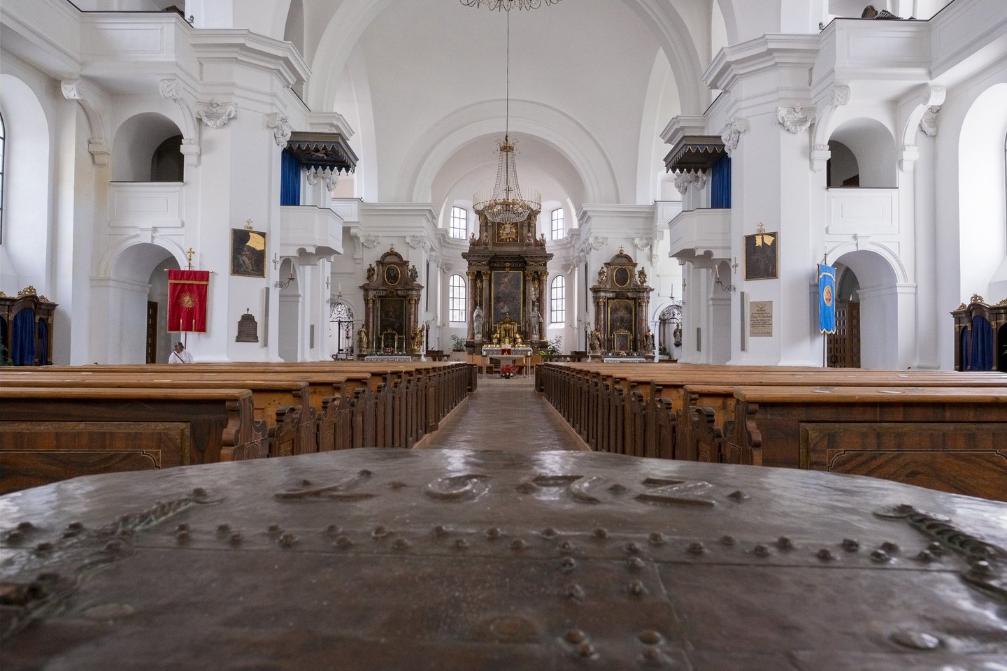 The white baroque nave of Hallein parish church seen down the central aisle from behind the studded metal lid of the baptismal font, with the gilded high altar at the far end.
