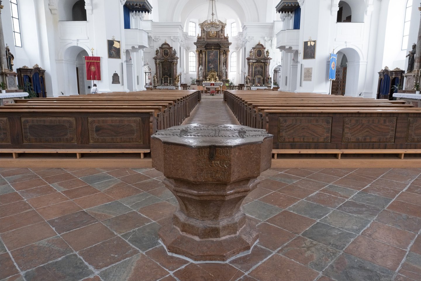 An octagonal red marble baptismal font with a hammered metal lid stands on the tiled floor of Hallein parish church, the pews and altar receding behind it.