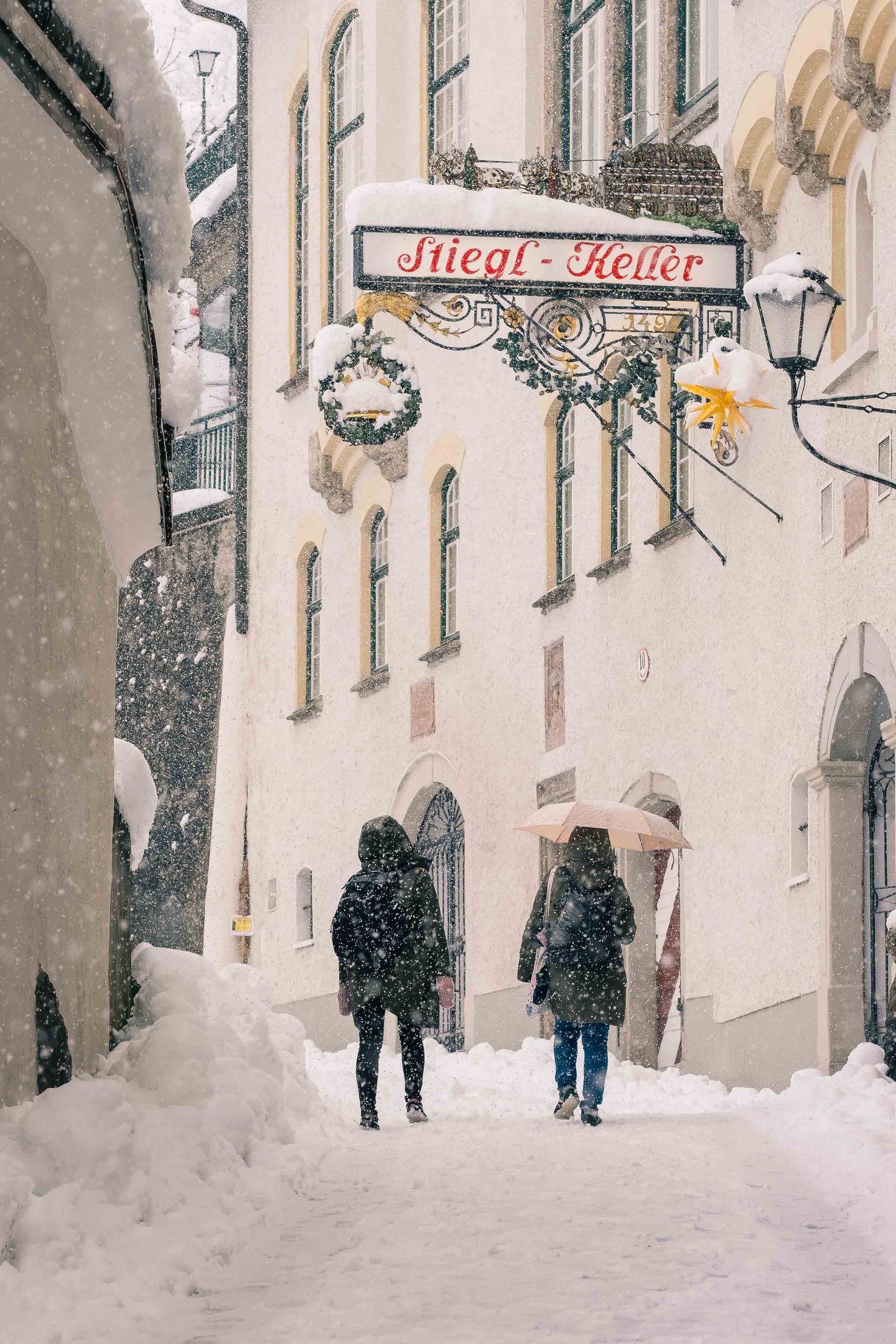 Two people walking toward Stieglkeller entrance under heavy snowfall in Salzburg old town.