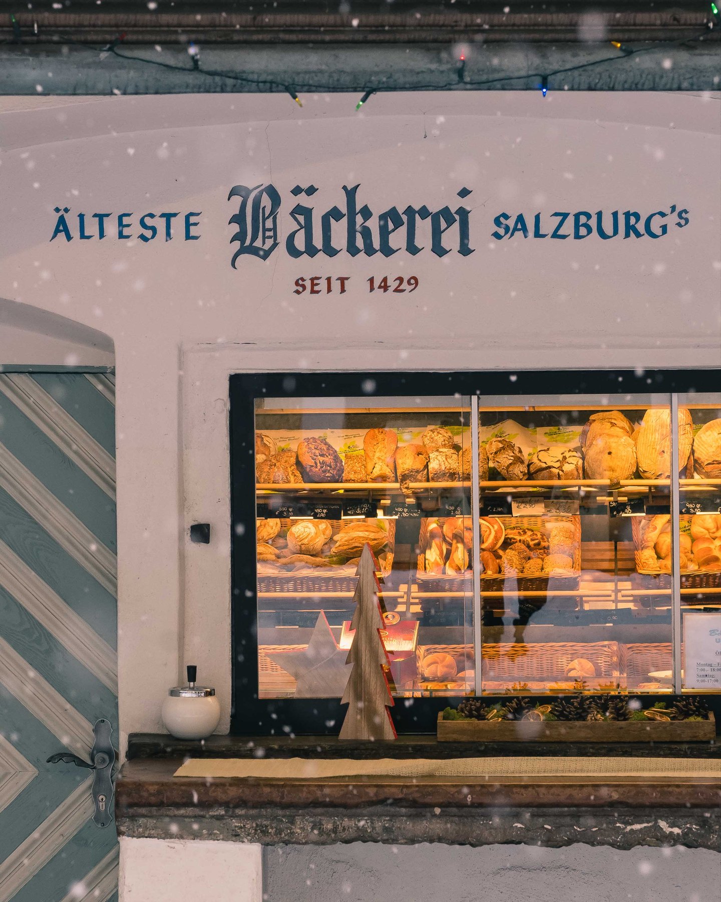 Snow falls past a Salzburg bakery storefront with Aelteste Baeckerei Salzburg's lettering above in winter