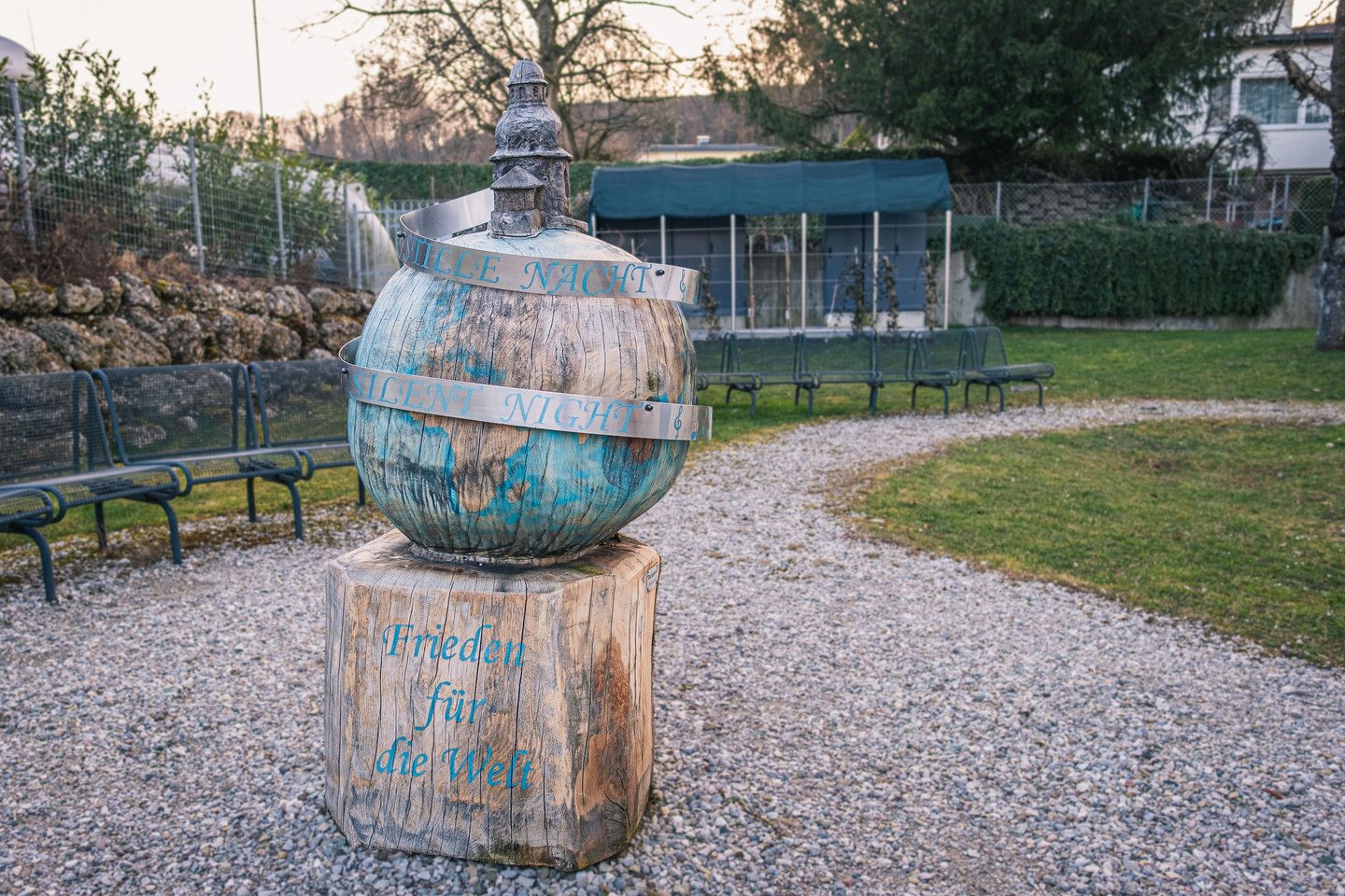 A carved wooden globe on a timber plinth in the grounds of the Silent Night Chapel in Oberndorf, banded with metal strips reading Stille Nacht and Silent Night above the words Frieden fuer die Welt.