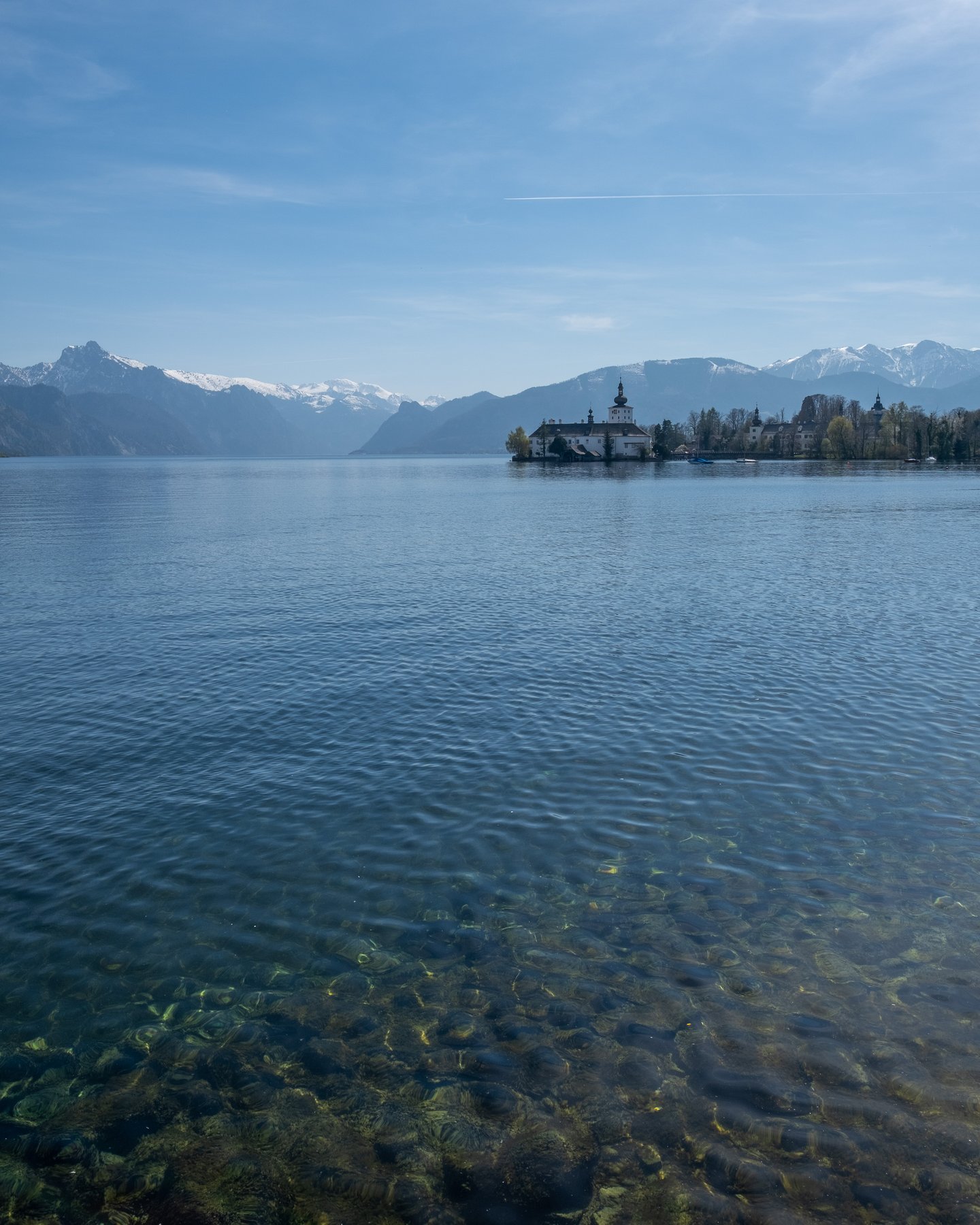 The Traunsee seen across clear stony shallows, with Seeschloss Ort on its island to the right and snow-capped Salzkammergut peaks on the far shore.
