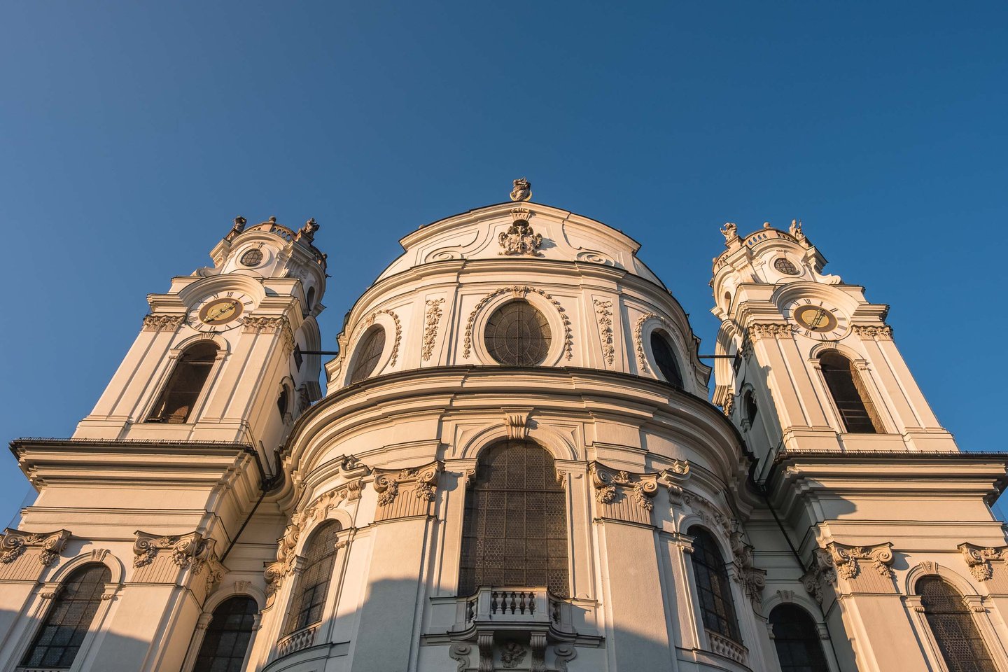 Baroque facade and twin towers of Kollegienkirche glow in golden hour light in Salzburg old town.