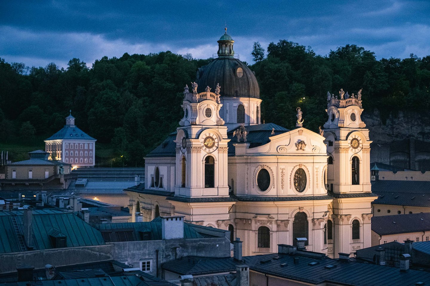 Illuminated Kollegienkirche facade glows above Altstadt rooftops at blue hour in Salzburg