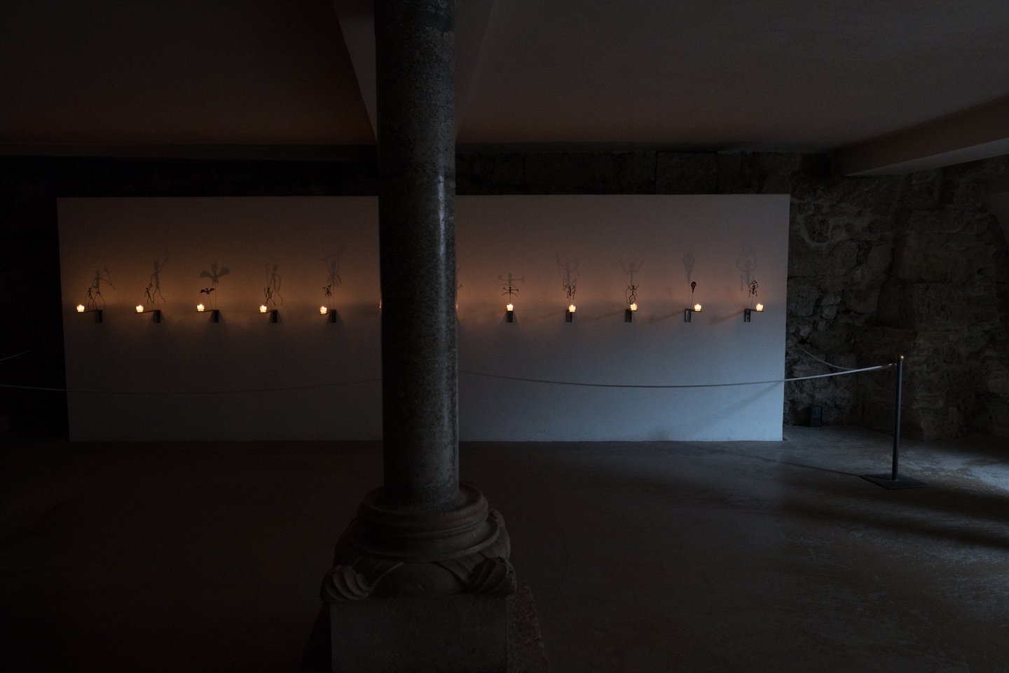 A wide dark view of the crypt beneath Salzburg Cathedral with a row of candle brackets and skeleton figures glowing along two pale panels behind a Romanesque column.