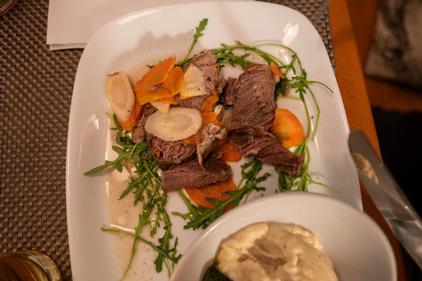 Seen from above, slices of boiled beef on a square white plate with carrot and parsnip discs and rocket, the bowl of creamed spinach and potato entering at the lower right.